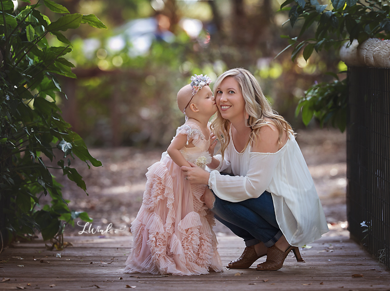 Pediatric leukemia girl in pink princess dress an floral crown kissing mom on the cheek on birdge